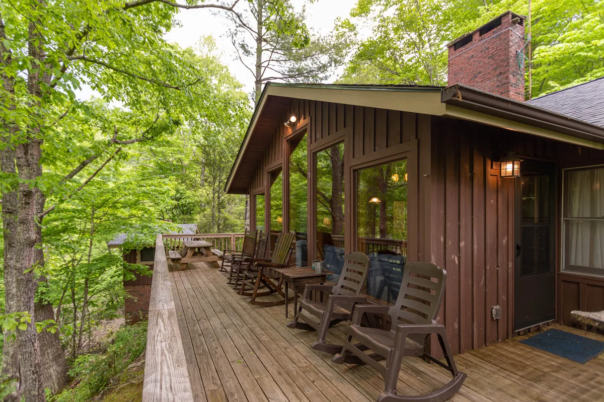 A spacious wooden deck set against a lush green backdrop, featuring several inviting rocking chairs arranged to take in the views. Large windows connect the outdoor space to the interior, and there is ample room for relaxing or socializing, suggesting a seamless indoor-outdoor lifestyle.