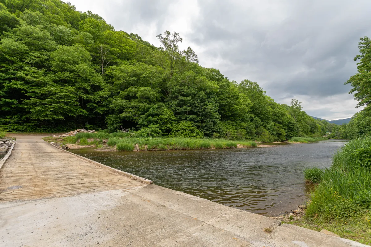 A serene view of the calm New River bordered by lush, tree-lined shores and a concrete pathway. The scene invites families to engage in riverside activities like fishing, hiking, and picnicking while soaking in the peaceful natural beauty.