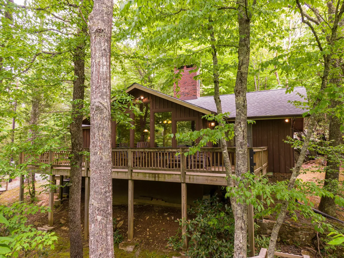 A wooden deck overlooks serene trees, with large windows reflecting the peaceful outdoors. Sunset hues create a cozy, intimate ambiance ideal for enjoying coffee or stargazing together.