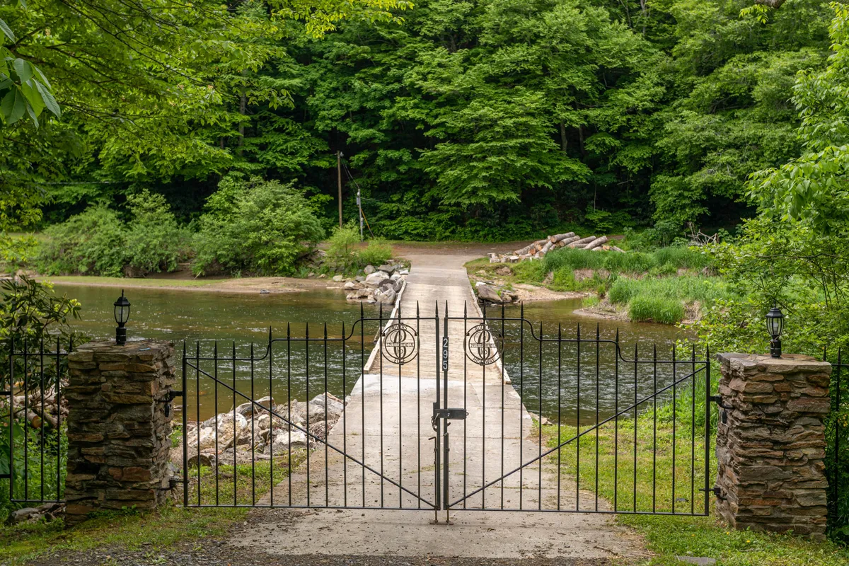 This image features an inviting iron gate opening to a tranquil pathway surrounded by lush trees, with glimpses of the adjacent water body. The sunset casts a warm glow over the scene, enhancing the natural beauty and sense of privacy.