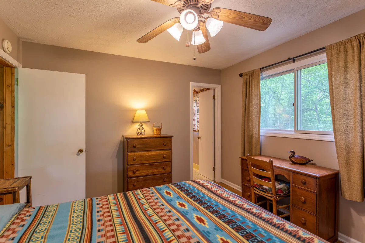 A warmly decorated bedroom featuring a colorful patterned bedspread, a wooden dresser, a desk and chair by a window with curtains. The room feels personal, comfortable, and functional, reflecting modern touches alongside homey charm.