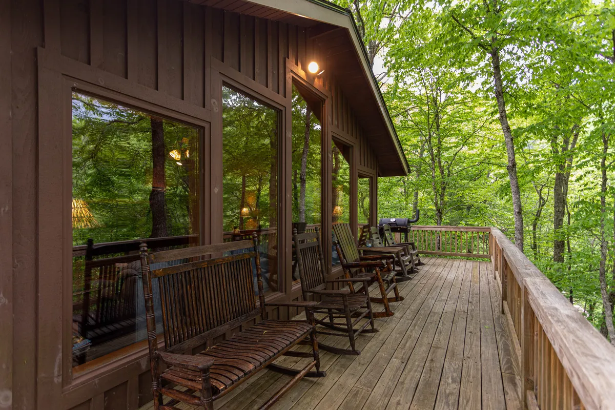 A cozy arrangement of rocking chairs lined up along a wooden deck, backed by large windows and surrounded by trees. The setting evokes a sense of relaxation and invites guests to sit, read, or simply watch the sunset and listen to the river.