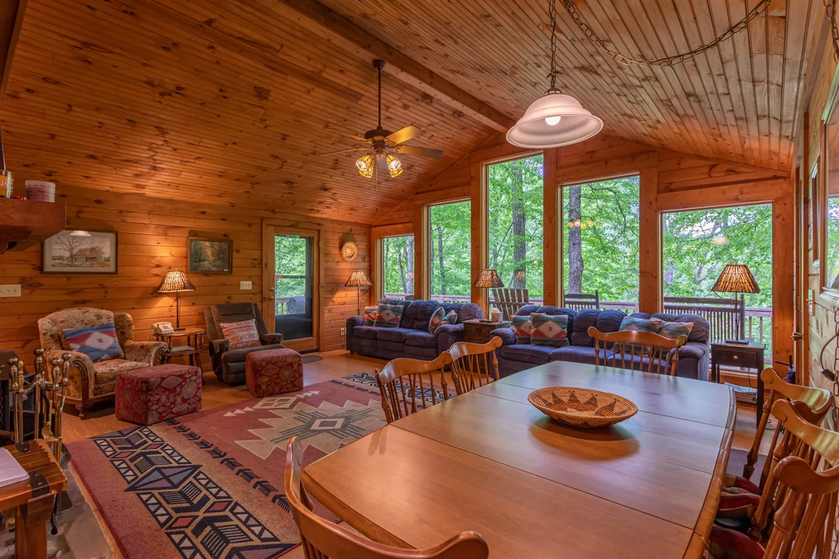 An open-concept living and dining area with a wooden ceiling, large windows that fill the space with natural light, a dining table, and a comfortable seating arrangement. The view through the windows suggests peaceful surroundings, ideal for shared meals and gatherings.