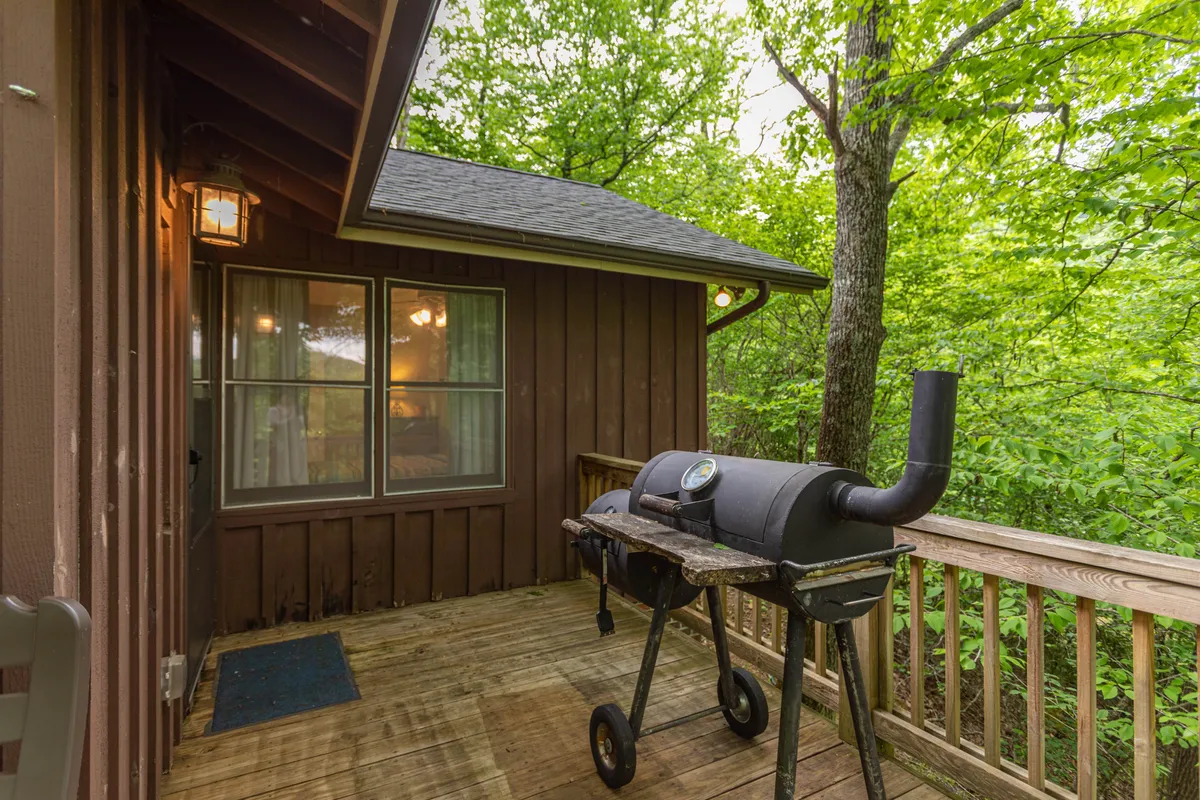A view of the wooden deck featuring a barbecue grill, surrounded by vibrant greenery and bordered by deck railings. The scene suggests space for outdoor cooking and gatherings, with windows nearby reflecting the natural surroundings.