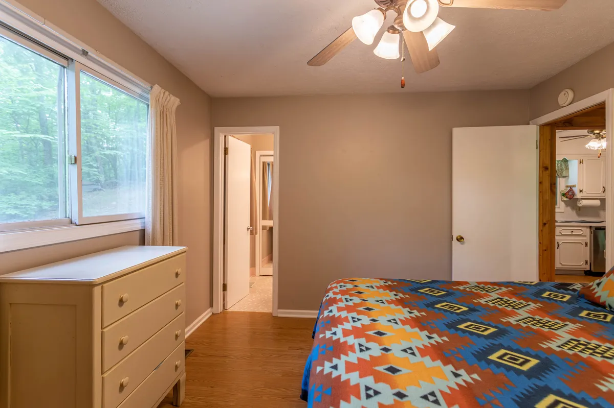 A bedroom with a colorful bedspread, a large window with serene forest views, and a dressing area doorway visible. The space feels bright, welcoming, and harmoniously connected to nature, perfect for waking up refreshed.