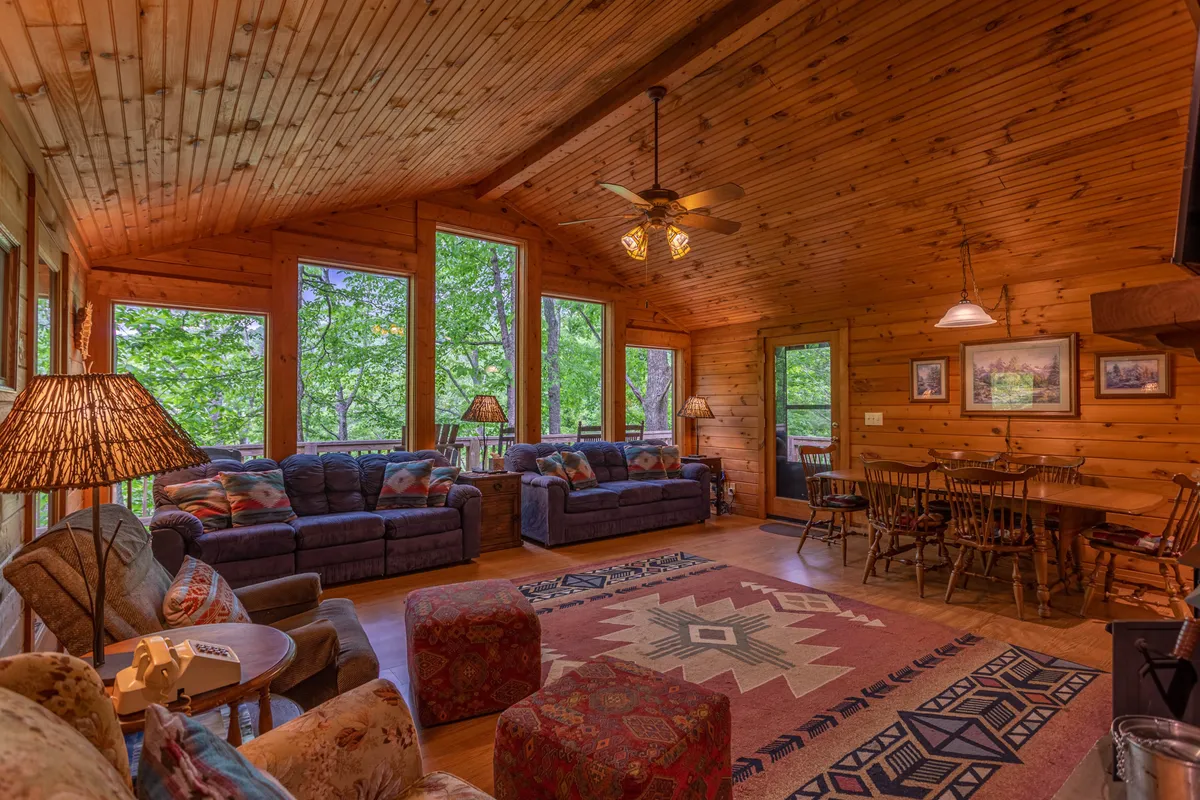 A spacious, welcoming great room filled with comfortable sofas, a decorative rug, and a dining table. Large windows offer expansive views of the surrounding trees and let in abundant natural light, creating a warm, inviting space perfect for gatherings. Table lamps add a cozy glow, and the layout flows naturally between the living and dining areas.