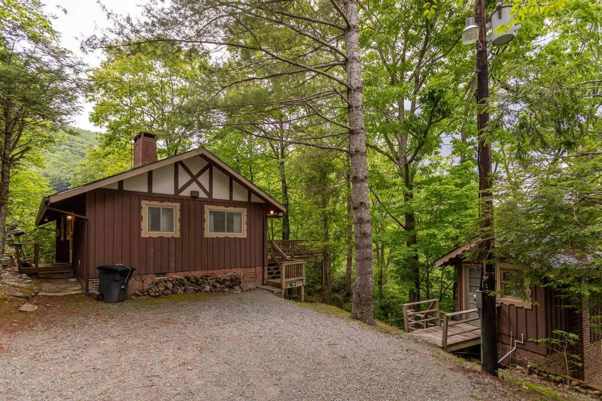A welcoming wooden cabin nestled among tall trees, with a gravel driveway leading up to the porch. The cabin is surrounded by natural greenery, with plenty of space for outdoor activities and relaxation.
