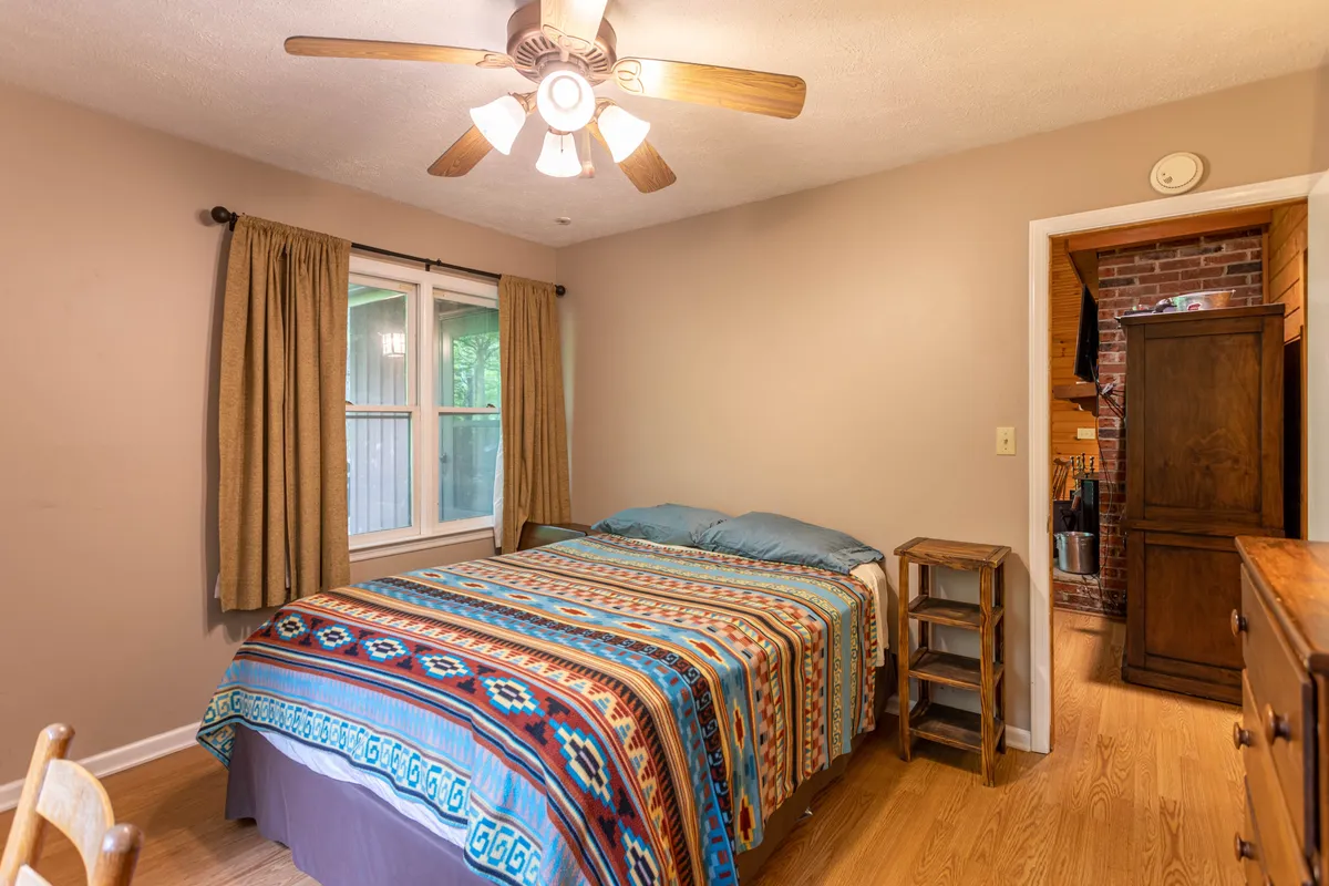 A bedroom showcasing a bed with a colorful blanket, a window framed by curtains, and wooden furniture that adds rustic charm. The setting is both inviting and intimate, suitable for quiet nights or sharing laughter with loved ones.